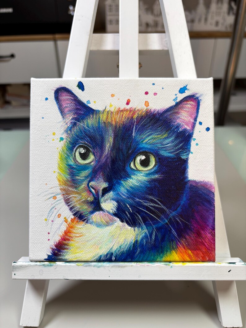 May include: A colorful painting of a cat's face on a white canvas. The cat has black fur with white markings and green eyes. The background is white with colorful paint splatters.