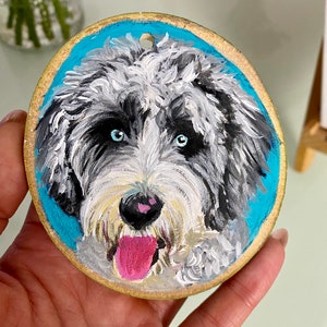 May include: A hand-painted wooden ornament featuring a portrait of a dog with white, black, and gray fur. The dog has blue eyes and a pink tongue sticking out. The background is a bright blue.