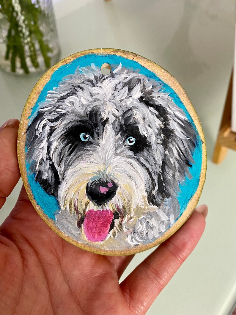 May include: A hand-painted wooden ornament featuring a portrait of a dog with white, black, and gray fur. The dog has blue eyes and a pink tongue sticking out. The background is a bright blue.