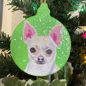 May include: A green glitter Christmas ornament with a painted image of a white chihuahua with pink ears.