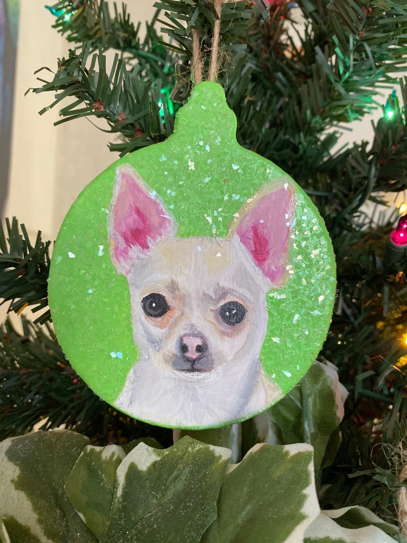 May include: A green glitter Christmas ornament with a painted image of a white chihuahua with pink ears.