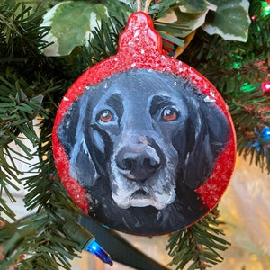 May include: A hand-painted Christmas ornament featuring a black Labrador Retriever's face on a red background with white flecks. The ornament is round and has a string for hanging.