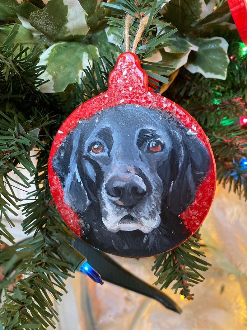 May include: A hand-painted Christmas ornament featuring a black Labrador Retriever's face on a red background with white flecks. The ornament is round and has a string for hanging.