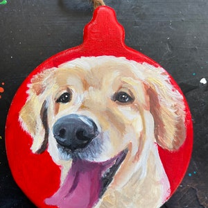 May include: A hand-painted Christmas ornament featuring a golden retriever dog with a pink tongue sticking out. The ornament is round and red with a brown twine hanger.