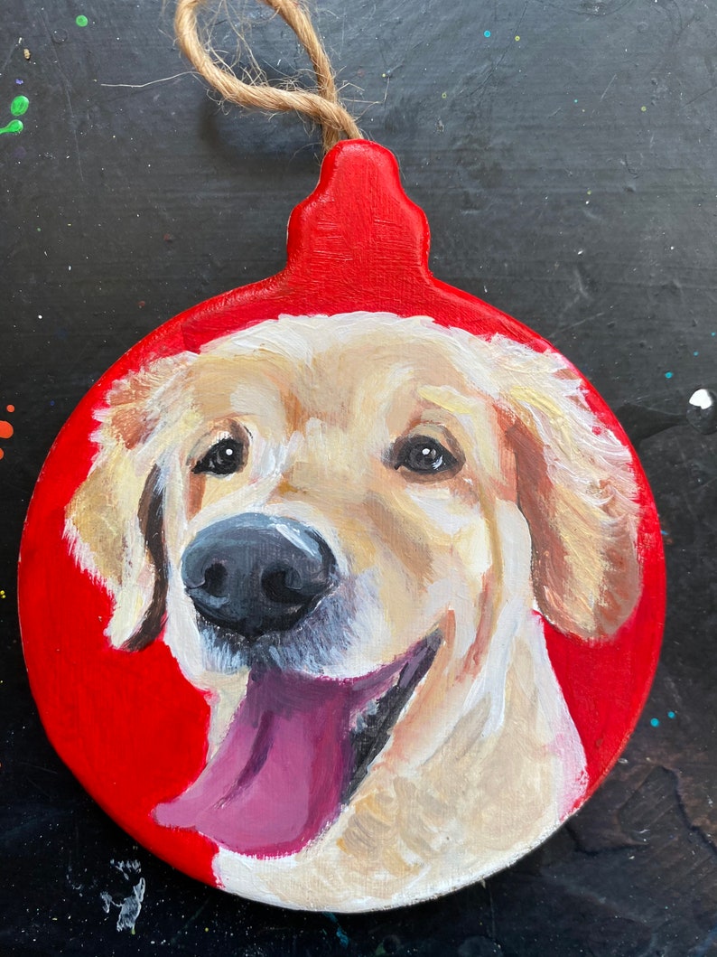 May include: A hand-painted Christmas ornament featuring a golden retriever dog with a pink tongue sticking out. The ornament is round and red with a brown twine hanger.
