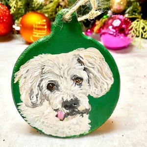 May include: A white dog with its tongue sticking out is painted on a green round Christmas ornament. The ornament has a twine hanger.