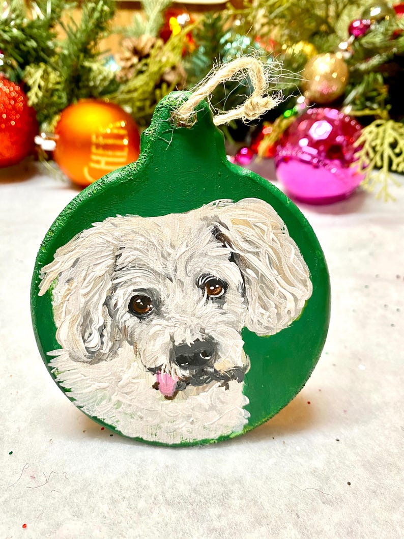 May include: A white dog with its tongue sticking out is painted on a green round Christmas ornament. The ornament has a twine hanger.