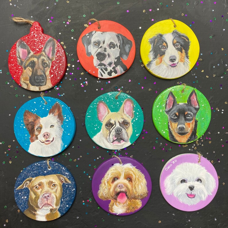 May include: Nine round Christmas ornaments with painted portraits of different dog breeds. The ornaments are painted on a white background with a variety of colors, including red, green, blue, purple, and yellow. The dogs are all smiling and looking at the camera.