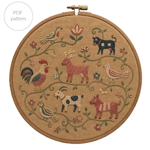May include: A circular wooden embroidery hoop containing a tan fabric panel with a PDF pattern label. The fabric features hand-stitched embroidery of a rooster, a deer, a goat, a dog, a cow, and three birds, all connected by green vines and flowers.