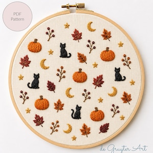 May include: A round wooden embroidery hoop containing a cream-colored fabric with hand-stitched motifs including orange pumpkins, black cats, red and brown autumn leaves, yellow crescent moons, small stars, and brown berry sprigs on stems.