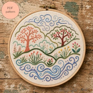May include: A circular wooden embroidery hoop containing a fabric panel with a hand-stitched landscape design. The scene features two trees, rolling green hills, colorful wildflowers, and swirling blue water patterns. Text reads PDF pattern.