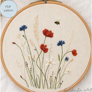 Könnte beinhalten: Ein runder Stickrahmen aus Holz mit einem Stoffeinsatz, der ein handgesticktes botanisches Motiv zeigt. Die Stickerei besteht aus roten Mohnblumen, blauen Kornblumen, weißen Wildblumen und hohen Gräsern, ergänzt durch eine kleine Biene.