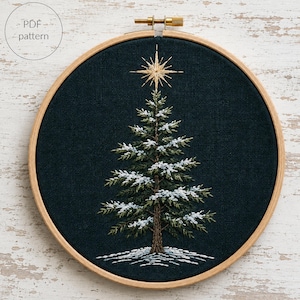 Peut inclure: Un tambour à broder circulaire en bois contenant un tissu bleu marine foncé avec un motif de sapin de Noël brodé à la main. L'arbre présente des branches vertes avec des accents de neige blanche, un tronc marron et une étoile dorée. Texte PDF pattern.
