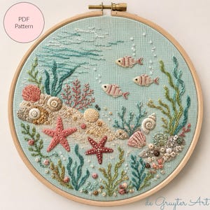Puede incluir: Un bastidor de bordado con tela azul claro que presenta una escena submarina bordada a mano con peces rosas, estrellas de mar, varias conchas y algas verdes. El diseño incluye pequeños nudos franceses blancos como burbujas.