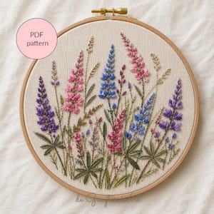 Puede incluir: Diseño floral bordado con flores de altramuces moradas, rosas y azules en un aro de madera. El diseño incluye hojas y tallos verdes. El texto "PDF pattern" está en un círculo rosa. Un proyecto de bordado artesanal.