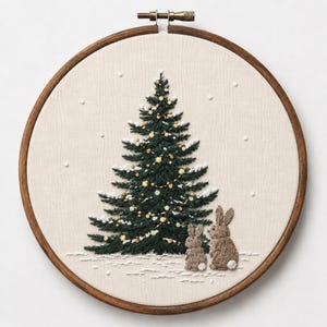 以下が含まれることがあります： 木製の円形刺繍枠に、手刺繍で施された濃い緑色のクリスマスツリーが描かれた布パネル。ツリーには金色のアクセントと白い雪が装飾されています。布地のツリーの根元には、2匹の小さな茶色のウサギが刺繍されています。