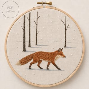 May include: A circular wooden embroidery hoop containing a fabric panel with a hand-stitched design of a red fox walking through a snowy forest. The scene features dark brown tree trunks, white French knot snowflakes, and the text PDF pattern.