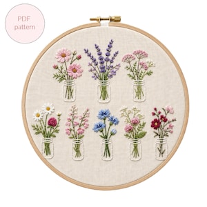 Puede incluir: Un bastidor de bordado que contiene un panel de tela beige con ocho arreglos florales bordados en pequeños frascos de vidrio. Las flores incluyen cosmos rosas, lavanda morada, acianos azules y varias flores silvestres en tonos rosa, blanco y rojo.