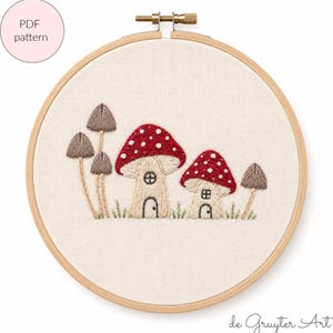 Può includere: Un telaio da ricamo contenente un tessuto color crema con un disegno ricamato a mano di due case a fungo con cappelli rossi a pois e tre funghi marroni più piccoli. Il testo PDF pattern è visibile nell'angolo in alto a sinistra.