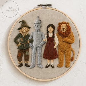 May include: A circular wooden embroidery hoop containing a hand-stitched fabric design of four characters: a scarecrow in green and brown, a silver tin person, a person in a brown dress, and a golden-brown lion. The text PDF Pattern is visible.