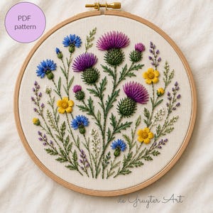 May include: A circular wooden embroidery hoop containing a fabric panel with a hand-stitched floral design featuring purple thistles, blue cornflowers, yellow buttercups, and small white and purple sprigs, with the text PDF pattern visible.