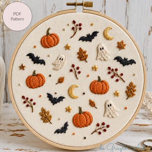 Könnte beinhalten: Ein Stickrahmen mit cremefarbenem Stoff, bestickt mit Motiven von orangefarbenen Kürbissen, weißen Geistern, schwarzen Fledermäusen, gelben Mondsicheln, braunen Herbstblättern und roten Beeren. Ein kreisförmiges Etikett trägt die Aufschrift PDF Pattern.