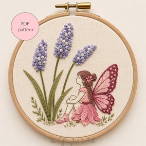 May include: Embroidered artwork in a wooden hoop. The design features a fairy with pink wings and dress, sitting near lavender flowers. The text "PDF pattern" is in a pink circle. The embroidery is on a cream-coloured fabric.