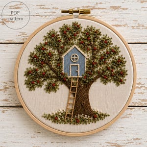 以下が含まれることがあります： 刺繍枠にはめられた布地に、緑の葉と赤い実をつけた大きな木の刺繍が施されています。枝の間には丸い窓とドアのある青い家があり、木製の梯子で地面と繋がっています。