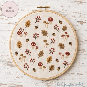 May include: An embroidery hoop containing off-white fabric with a pattern of stitched autumn elements including red and white mushrooms, brown oak leaves, small acorns, and clusters of red berries. The text PDF Pattern is in the top left corner.