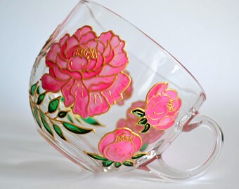 May include: A clear glass teacup featuring hand-painted pink peony flowers with gold outlines and green leaves. The design wraps around the exterior of the cup, which has a curved body and a side handle, showcasing vibrant floral artwork.
