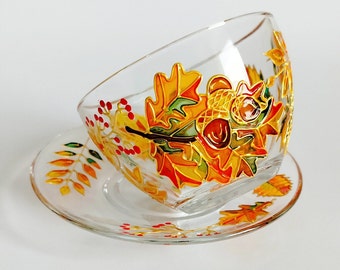 May include: A clear glass teacup and saucer set featuring hand-painted autumn motifs. The design includes vibrant orange, yellow, and green oak leaves, brown acorns, and small red berries outlined in gold, decorating both the cup and the saucer.