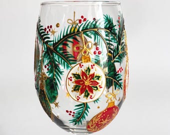 May include: A clear glass stemware piece featuring hand-painted holiday decorations. The design includes green pine branches, red berries, and gold-accented Christmas ornaments, with one central white ornament displaying a red poinsettia flower.