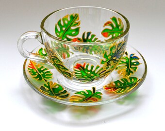 May include: A clear glass teacup and matching saucer set featuring a hand-painted pattern of vibrant green monstera leaves with yellow and orange accents outlined in gold. The cup has a curved handle and a circular base, while the saucer is round.