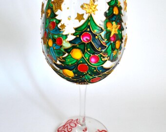 May include: A clear glass wine glass featuring hand-painted green Christmas trees decorated with red and yellow ornaments and gold stars. The base of the glass is painted with the red text Cheers and the year 2026 in a script font.