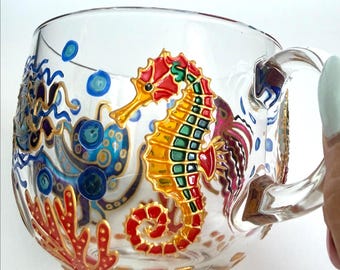 May include: A clear glass mug featuring a hand-painted design of a vibrant red and yellow seahorse, a blue octopus, and red coral. The artwork includes small blue bubbles and gold outlines, creating a detailed underwater scene on the glass surface.