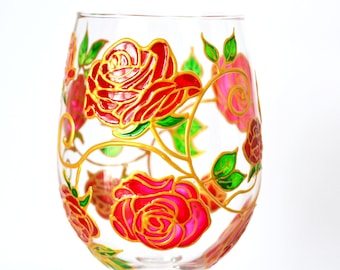 May include: A clear glass wine glass decorated with hand-painted red and pink roses featuring gold outlines. The design includes winding gold vines and vibrant green leaves that wrap around the exterior surface of the glass bowl.