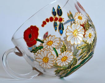 May include: A clear glass teacup featuring hand-painted floral designs including white daisies with yellow centers and a single red poppy. A colorful dragonfly with red, blue, and gold accents is painted above the flowers. The cup has a handle.
