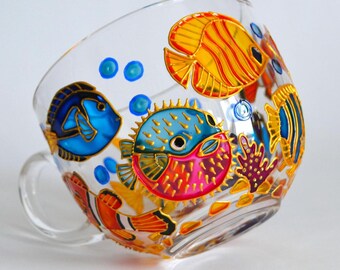 May include: A clear glass mug decorated with hand-painted colorful marine life, including a bright blue pufferfish, a yellow angelfish, and an orange clownfish, surrounded by small blue bubbles and purple coral accents on the exterior surface.