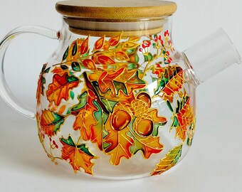 May include: A clear glass teapot featuring a hand-painted design of vibrant autumn leaves, acorns, and small red berries. The teapot has a rounded body, a curved glass handle, a short spout, and a circular light brown wooden lid with a flat top.