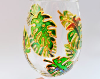 May include: A clear glass wine glass featuring hand-painted monstera leaves in vibrant shades of green, yellow, and orange. The leaves are outlined with a raised gold-toned paint, creating a textured effect across the surface of the glass.