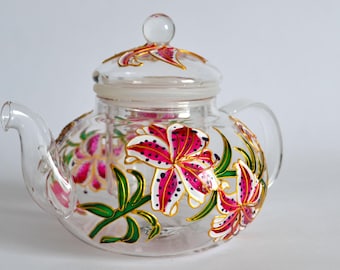 May include: A clear glass teapot featuring a hand-painted design of vibrant pink and white lilies with green leaves and gold outlines. The teapot includes a matching glass lid with a spherical knob and a curved handle and spout.