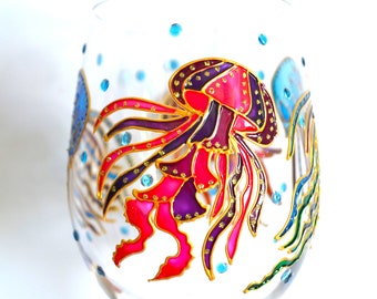 May include: A clear glass stemless wine glass featuring hand-painted jellyfish designs in vibrant shades of pink, purple, and blue. The jellyfish are outlined in gold paint with small gold dots, accented by scattered blue dots across the glass surface.