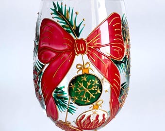 May include: A clear glass stemless wine glass featuring a hand-painted design of a large red bow tied around green pine branches. Below the bow, a green circular ornament with a gold snowflake pattern and a smaller red ornament are painted.