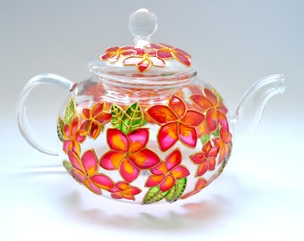 May include: A clear glass teapot featuring a hand-painted floral design with vibrant pink, orange, and yellow petals outlined in gold. The teapot includes a matching glass lid with a knob handle and a curved spout, decorated with green leaves.