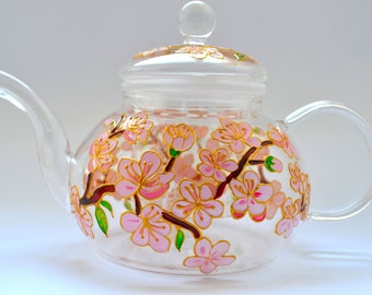 May include: A clear glass teapot featuring a hand-painted design of pink cherry blossoms on brown branches with green leaves. The teapot includes a matching glass lid with a spherical knob, a curved spout, and a rounded handle on the side.