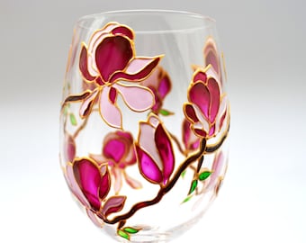 May include: A clear glass stemless wine glass decorated with hand-painted magnolia flowers in shades of deep magenta and light pink. The floral design features dark brown branches and small green leaves outlined in gold, wrapping around the glass.