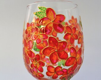 May include: A clear glass wine glass decorated with hand-painted plumeria flowers in vibrant shades of red, orange, and pink. The floral design features gold-colored outlines and small green leaves, wrapping around the exterior of the glass bowl.