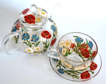 May include: A clear glass teapot with a matching lid, teacup, and saucer, all hand-painted with vibrant red poppies, blue cornflowers, and white daisies. The floral designs feature gold-toned outlines, creating a detailed botanical pattern.