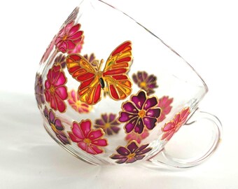 May include: A clear glass teacup featuring hand-painted designs of a vibrant orange and yellow butterfly surrounded by multiple pink and purple flowers. The floral and butterfly motifs are outlined in gold, creating a decorative pattern across the exterior surface of the rounded glass cup with a handle.