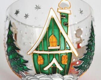 May include: A clear glass bowl featuring a hand-painted winter scene with a green house, a chimney with white smoke, and two green pine trees. The design includes yellow windows, white snow-covered ground, and silver stars against a snowy backdrop.